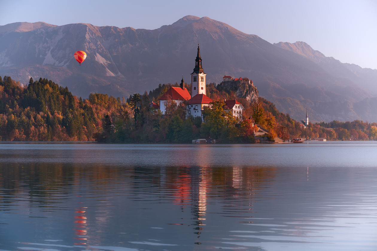 Church on a lake island below autumn mountains with a red hot-air balloon in the sky